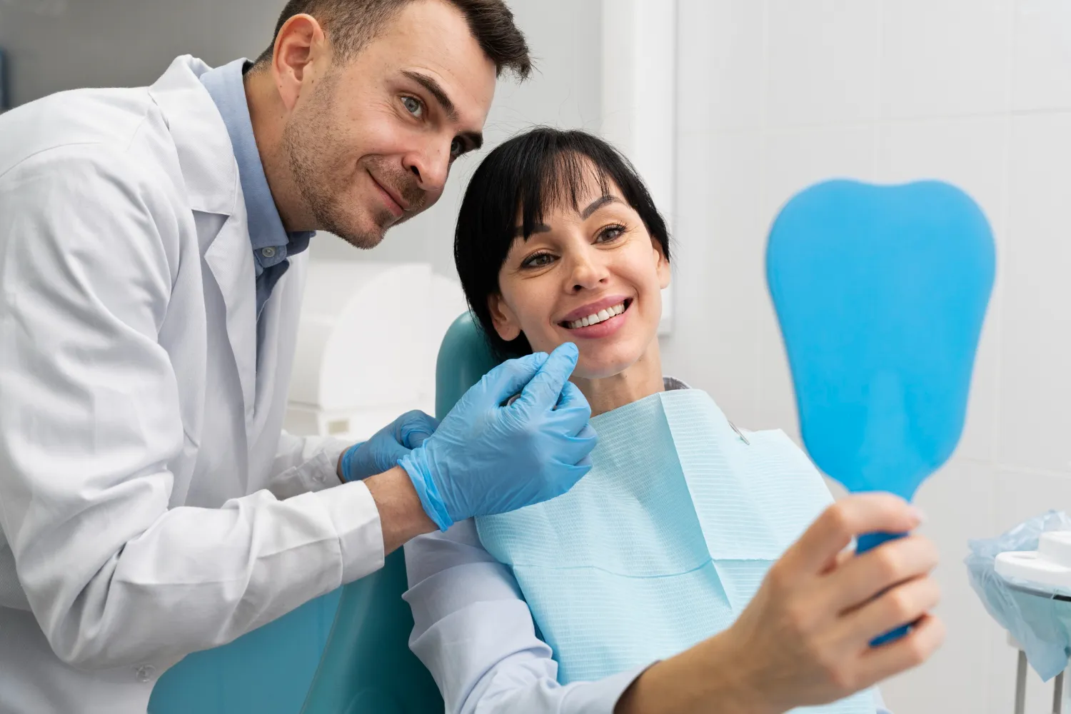 A dentist in a white coat examines a patient seated in a dental chair, while the patient holds a blue mirror, wearing a bib.
